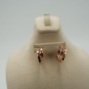 Peut inclure: Une paire de boucles d'oreilles créoles en or rose avec un design en forme de serpent. Les boucles d'oreilles sont composées de petits segments rectangulaires et ornées de petites pierres précieuses roses. Elles sont présentées sur un mannequin de couleur neutre.