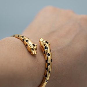 Puede incluir: Una pulsera abierta de tono dorado con dos cabezas de leopardo estilizadas en los extremos. La superficie metálica pulida está decorada con manchas de esmalte negro que imitan el patrón del pelaje de un leopardo a lo largo de la banda.