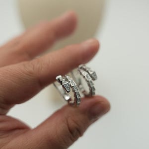 Può includere: Un anello color argento a forma di serpente avvolto, caratterizzato da un motivo a scaglie testurizzato lungo la fascia e piccole pietre chiare e scintillanti incastonate sulla superficie superiore della testa e del corpo del serpente.