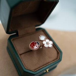 May include: A rose gold ring set featuring a ladybug design with red enamel and black spots, and a flower design with white mother-of-pearl petals. The rings are displayed in a green jewelry box with a brown interior.