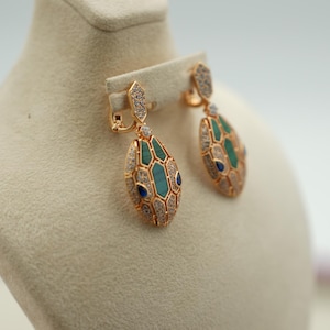 May include: Gold-toned drop earrings with a snake head design. The earrings feature emerald green and blue accents, with clear crystals and a detailed, textured pattern. The earrings are displayed on a cream-colored mannequin.