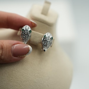 May include: Silver-toned snake head stud earrings with green gemstone eyes and clear crystal accents. The earrings are displayed on a neutral-toned surface, showcasing their intricate design and sparkling details. These earrings are a unique jewelry item.