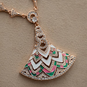Può includere: Collana color oro rosa con un ciondolo a forma di ventaglio. Il ciondolo presenta dettagli in smalto bianco, rosa e verde, bordati da cristalli trasparenti. La collana ha una catena delicata con abbellimenti di cristallo.
