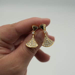 Könnte beinhalten: Ein Paar goldfarbene Ohrringe mit fächerförmigem Design. Die Ohrringe sind mit kleinen, klaren Edelsteinen besetzt, die einen funkelnden Effekt erzeugen. Die Ohrringe werden vor einem schlichten weißen Hintergrund gehalten.