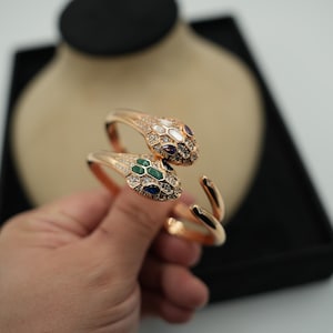 Puede incluir: Pulsera de serpiente en oro rosa con dos cabezas de serpiente. Las cabezas de serpiente están adornadas con piedras preciosas blancas, verdes y moradas. La pulsera se muestra sobre un fondo neutro.