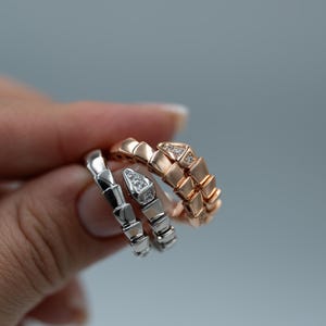 Pode incluir: Três anéis em forma de cobra em prata e ouro rosa. O anel de ouro rosa tem um detalhe de diamante. Os anéis são segurados em uma mão contra um fundo cinza. Os anéis são uma joia.