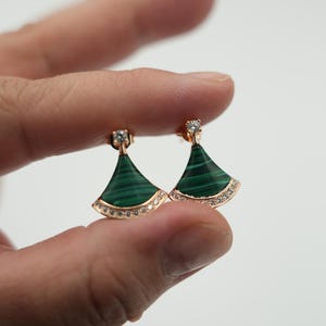 May include: Gold-coloured earrings with a fan-shaped design. The earrings have a vibrant green malachite inlay and are embellished with small, sparkling clear stones. The earrings are held up by a person's fingers.