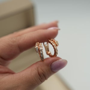 May include: Rose gold hoop earrings in a snake design, adorned with small, sparkling gemstones. The earrings are displayed against a neutral backdrop, highlighting their intricate details and elegant style.