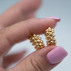 May include: A pair of gold hoop earrings featuring a textured surface with repeating pyramid-shaped studs and small spherical accents along the outer edge. The earrings have a polished metallic finish and a circular design.