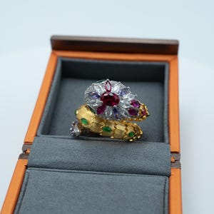 May include: A gold-toned snake-shaped ring featuring a textured scale pattern with small green accents. The head of the snake is adorned with a cluster of clear, purple, and red marquise-cut stones, centred by a prominent oval red gemstone.