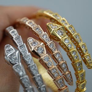 Op de afbeelding: Een verzameling slangenarmbanden in goud, roségoud en zilver, elk versierd met sprankelende, heldere edelstenen. De armbanden worden getoond tegen een zachte, neutrale achtergrond, die hun ontwerp benadrukt.