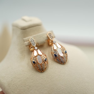Peut inclure: Boucles d'oreilles dorées en forme de serpent avec des accents blancs, bleus et transparents. Les boucles d'oreilles présentent un design pendant avec une surface détaillée et texturée. La tête du serpent est ornée de pierres précieuses et d'incrustations de nacre.