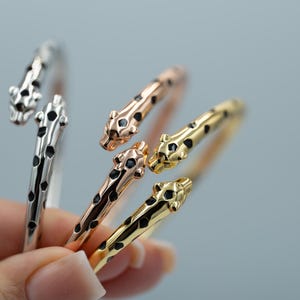 May include: Three open-ended cuff bracelets shaped like panther heads, featuring black enamel spots. One bracelet is silver-toned, one is rose gold-toned, and one is gold-toned. Each piece has a stylized feline head at both ends of the open band.