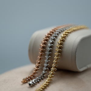Puede incluir: Tres pulseras metálicas en tonos oro rosa, plata y oro amarillo. Cada pulsera presenta una hilera continua de pequeñas púas puntiagudas que se alternan con diminutas piedras transparentes de corte redondo, creando un aspecto texturizado.