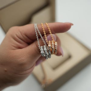 Könnte beinhalten: Drei Halsketten mit Anhängerringen, eine silberne, eine goldene und eine roségoldene, jeweils mit einer Kette. Die Ringe sind mit kleinen, funkelnden Steinen besetzt. Die Halsketten werden vor einer neutralfarbenen Box präsentiert.