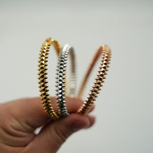 Puede incluir: Tres brazaletes circulares con una superficie texturizada de púas. El conjunto incluye un brazalete de metal en tono dorado, uno plateado y uno en tono oro rosa, cada uno con un patrón repetitivo de pequeños tachones puntiagudos en el borde.