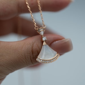 May include: Rose gold necklace with a fan-shaped pendant. The pendant has a white, pearlescent centre and is outlined with small, clear gemstones. A single gemstone accents the centre of the pendant. The necklace is held by a hand against a light background.