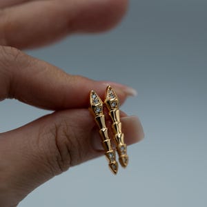 Könnte beinhalten: Ein Paar goldfarbene Ohrringe mit einem segmentierten Design, jedes Segment mit kleinen, klaren Edelsteinen besetzt. Die Ohrringe werden vor einem hellgrauen Hintergrund gehalten.