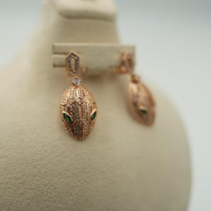 Può includere: Un paio di orecchini pendenti in oro rosa a forma di testa di serpente, caratterizzati da un motivo a scaglie testurizzato incastonato con piccole pietre chiare e due piccole pietre verdi come occhi. Gli orecchini pendono da un anello geometrico.