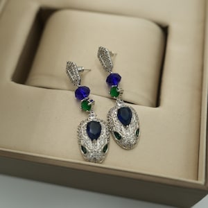 Könnte beinhalten: Silberfarbene Ohrhänger mit Schlangenmuster. Die Ohrringe haben einen großen, tropfenförmigen blauen Edelstein, mit kleineren blauen und grünen Edelsteinen darüber. Die Schlangenköpfe sind mit kleinen, klaren Kristallen besetzt.
