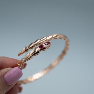 May include: Rose gold snake bracelet with a coiled design. The bracelet features a snake head with two red gemstones for eyes. The bracelet is a stylish piece of jewellery.