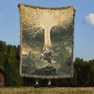Puede incluir: Una manta tejida con una ilustración detallada de un gran árbol y un caballero a caballo. El diseño es en tonos beige, gris y negro, con un borde con flecos. La manta se sostiene contra un cielo azul.