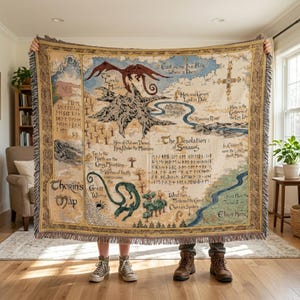 Puede incluir: Una manta de tapiz con un mapa con texto e ilustraciones. El mapa representa un paisaje de fantasía con montañas, ríos y criaturas. La paleta de colores incluye beige, marrón, azul y verde.