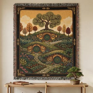 Puede incluir: Tapiz tejido que representa un paisaje caprichoso con casas de hobbits en una ladera verde. La obra de arte incluye un gran árbol, flores coloridas y un camino de piedra. El tapiz tiene un borde negro y bordes con flecos.