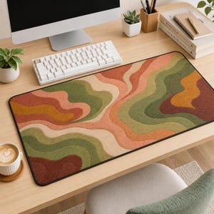 May include: A rectangular desk mat featuring a wavy, layered pattern in shades of olive green, terracotta, mustard yellow, and cream. The mat has a black stitched border and is positioned on a wooden desk surface with a computer and accessories.