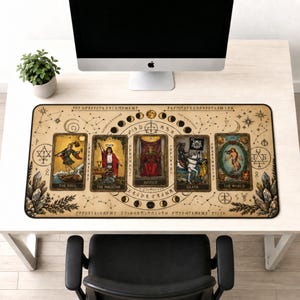 Puede incluir: Una alfombrilla de escritorio rectangular beige con ilustraciones de cartas del tarot. La alfombrilla muestra cinco cartas: El Loco, El Mago, Justicia, Muerte y El Mundo. El diseño incluye constelaciones, runas y fases lunares.