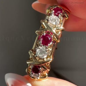 May include: A gold ring featuring an alternating pattern of three round red rubies and two round clear diamonds. The stones are set within a decorative gold band characterized by an intricate, crisscrossing metal design that wraps around each gem.