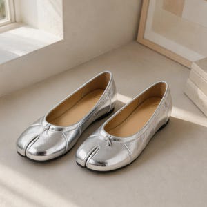May include: A pair of silver ballet flats with a split-toe design. The shoes have a shiny metallic finish, a small bow detail on the vamp, and a light brown insole. The flats are displayed on a neutral surface.