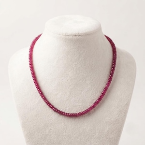 May include: A necklace composed of numerous small, round, faceted deep pinkish-red beads strung together in a continuous circular strand. The beads exhibit a uniform size and shape, creating a consistent texture throughout the entire length of the piece.