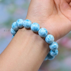 May include: A light blue beaded bracelet with a marbled effect. The bracelet, made of round beads, is worn on a wrist. The image includes the text "Radhika Gems Creations".