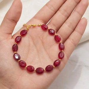 Könnte beinhalten: Ein Armband mit glatten, ovalen, tiefroten Perlen, die sich mit kleinen, runden, goldfarbenen Metallzwischenstücken abwechseln. Das Schmuckstück verfügt über einen goldfarbenen Metallkettenabschnitt mit einem Verschluss, der ein kreisförmiges Design bildet.