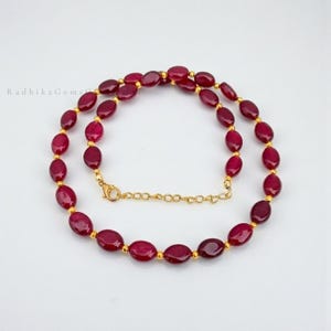 May include: A necklace featuring oval-shaped deep red ruby beads alternating with small round gold-toned metal beads. The piece includes a gold-toned metal lobster claw clasp and an adjustable chain link extender for a secure fit around the neck.