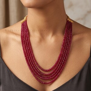 May include: A multi-strand necklace featuring five rows of small, faceted, deep red beads. The strands are gathered at the top by gold-toned metal caps with textured detailing. The text Radhika Creations is visible on the left side of the image.