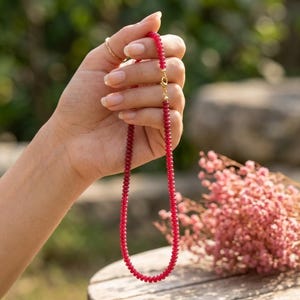 May include: A necklace composed of small, round, translucent red beads strung together. The necklace features a gold-toned metal lobster claw clasp at the top, connecting the two ends of the beaded strand into a circular loop.