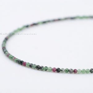 May include: A single strand of small, faceted, round beads in shades of green, black, and magenta. The beads are arranged in a curved line against a plain white background. The text RadhikaGemsCreations is visible on the left side of the image.