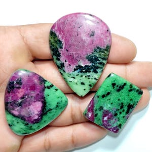 May include: Three polished zoisite gemstones featuring vibrant magenta ruby inclusions and deep green zoisite matrix. The stones are shaped as a teardrop, a triangle, and a rectangle, each displaying unique natural color patterns and smooth surfaces.