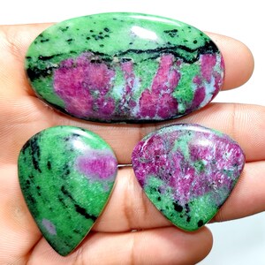 Peut inclure: Trois pierres précieuses en zoïsite polie présentant des inclusions de rubis magenta vif et des stries noires de hornblende. Une grande pierre ovale est placée au-dessus de deux pierres en forme de goutte, toutes avec une finition lisse.