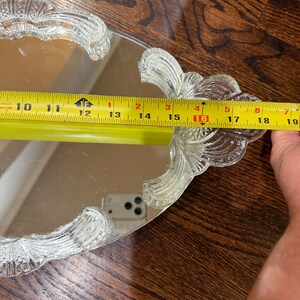 May include: An oval mirrored vanity tray with clear, textured glass handles featuring a fan-like pattern. A yellow measuring tape is placed across the tray, indicating a width of approximately 19 cm between the two glass handles.