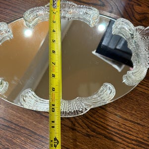 May include: An oval mirrored vanity tray with a clear, pressed glass border featuring decorative, textured handles. A yellow Stanley measuring tape is placed across the centre, indicating the tray width is approximately 26.7 centimetres.