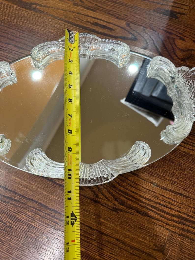 May include: An oval mirrored vanity tray with a clear, pressed glass border featuring decorative, textured handles. A yellow Stanley measuring tape is placed across the centre, indicating the tray width is approximately 26.7 centimetres.