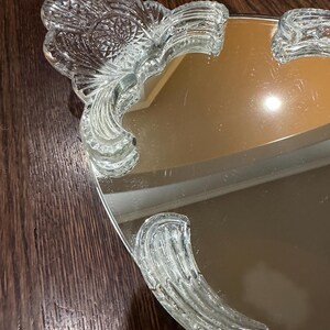 May include: A vanity tray featuring a reflective mirrored surface and clear glass handles. The handles exhibit a pressed glass pattern with fan-shaped motifs and intricate geometric detailing along the edges, providing a decorative border.