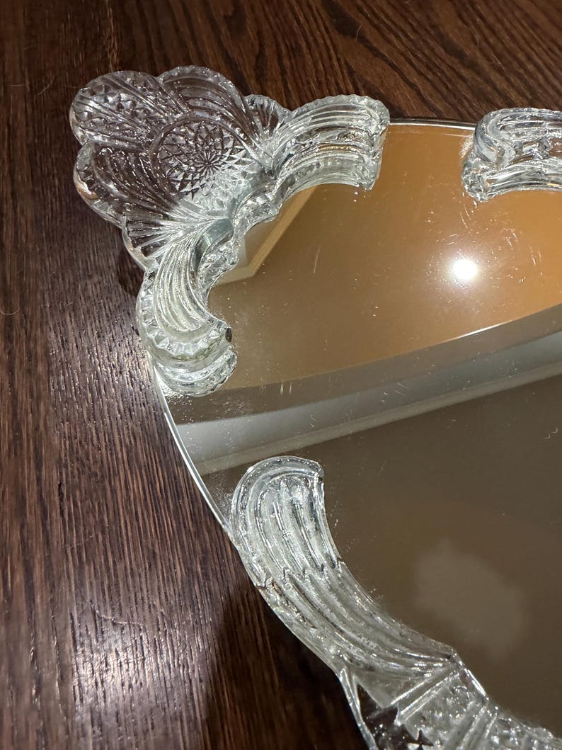 May include: A vanity tray featuring a reflective mirrored surface and clear glass handles. The handles exhibit a pressed glass pattern with fan-shaped motifs and intricate geometric detailing along the edges, providing a decorative border.