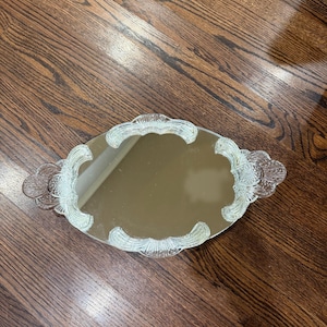 May include: An oval-shaped vanity tray featuring a central mirrored surface surrounded by a clear, textured glass border. The glass frame displays a repeating scalloped pattern with integrated handles on both sides, creating a decorative edge.