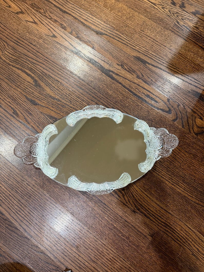 May include: An oval-shaped vanity tray featuring a central mirrored surface surrounded by a clear, textured glass border. The glass frame displays a repeating scalloped pattern with integrated handles on both sides, creating a decorative edge.