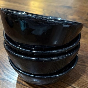May include: A stack of four glossy black ceramic bowls with a smooth, rounded design. Each bowl features a deep, uniform black finish with a reflective surface, stacked vertically to show the consistent shape and dark color of the set.