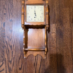 May include: A wooden wall clock with a shelf. The clock has a square face with Roman numerals and decorative floral accents. The clock and shelf are made of light brown wood. The clock is approximately 12 inches tall.